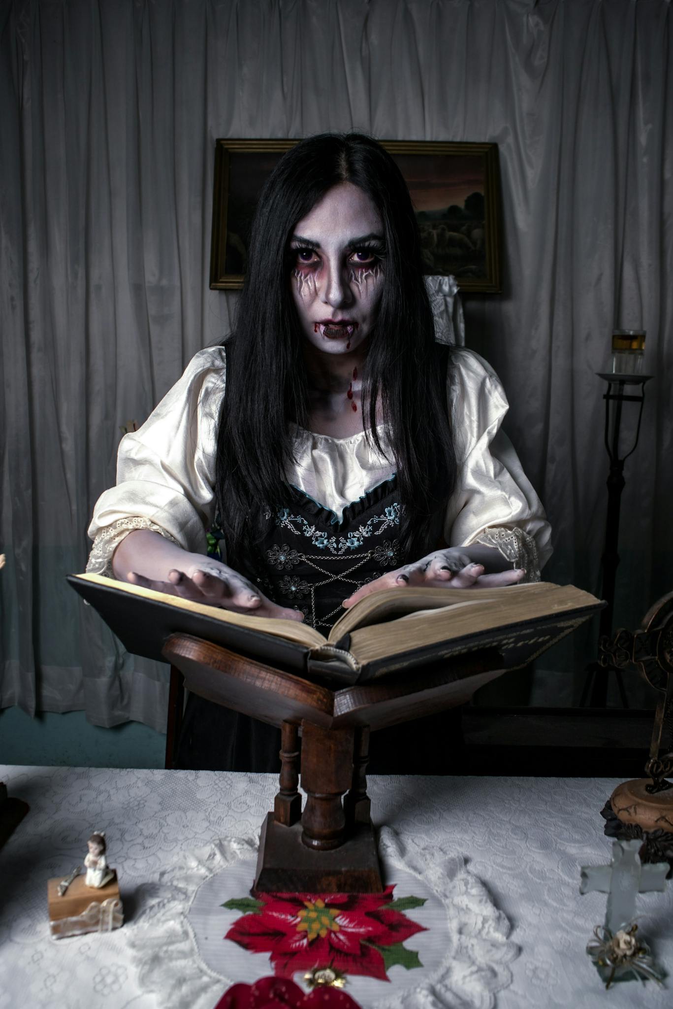 A mysterious woman in gothic attire stands reading a spellbook in a dimly lit room, creating an eerie atmosphere.