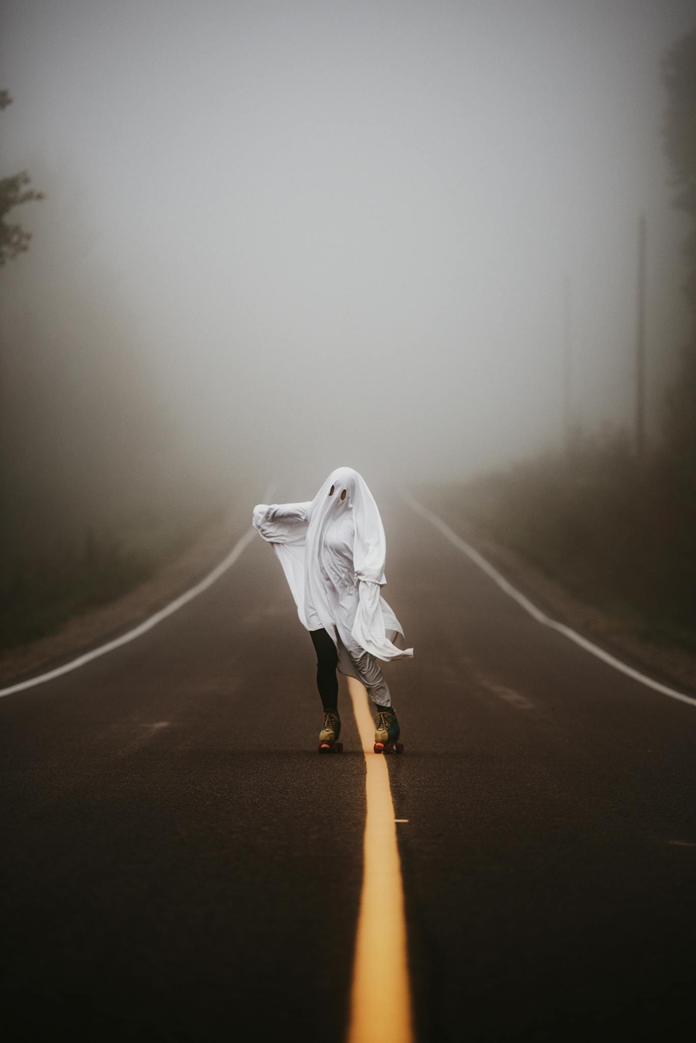 A person in a ghost costume roller skating on a foggy country road at dawn, creating an eerie scene.