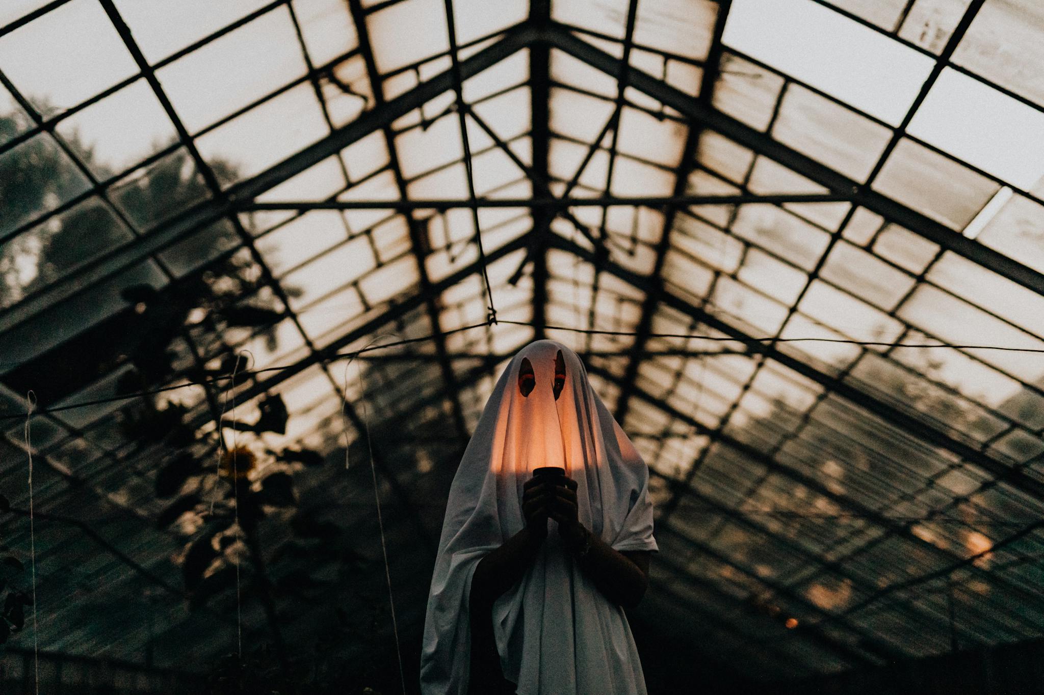 Mysterious figure in ghost costume holding lantern inside a dimly lit greenhouse.